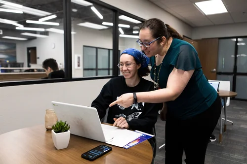 A mentor leans in to help a student review information on a laptop during a collaborative study session in a modern workspace.
